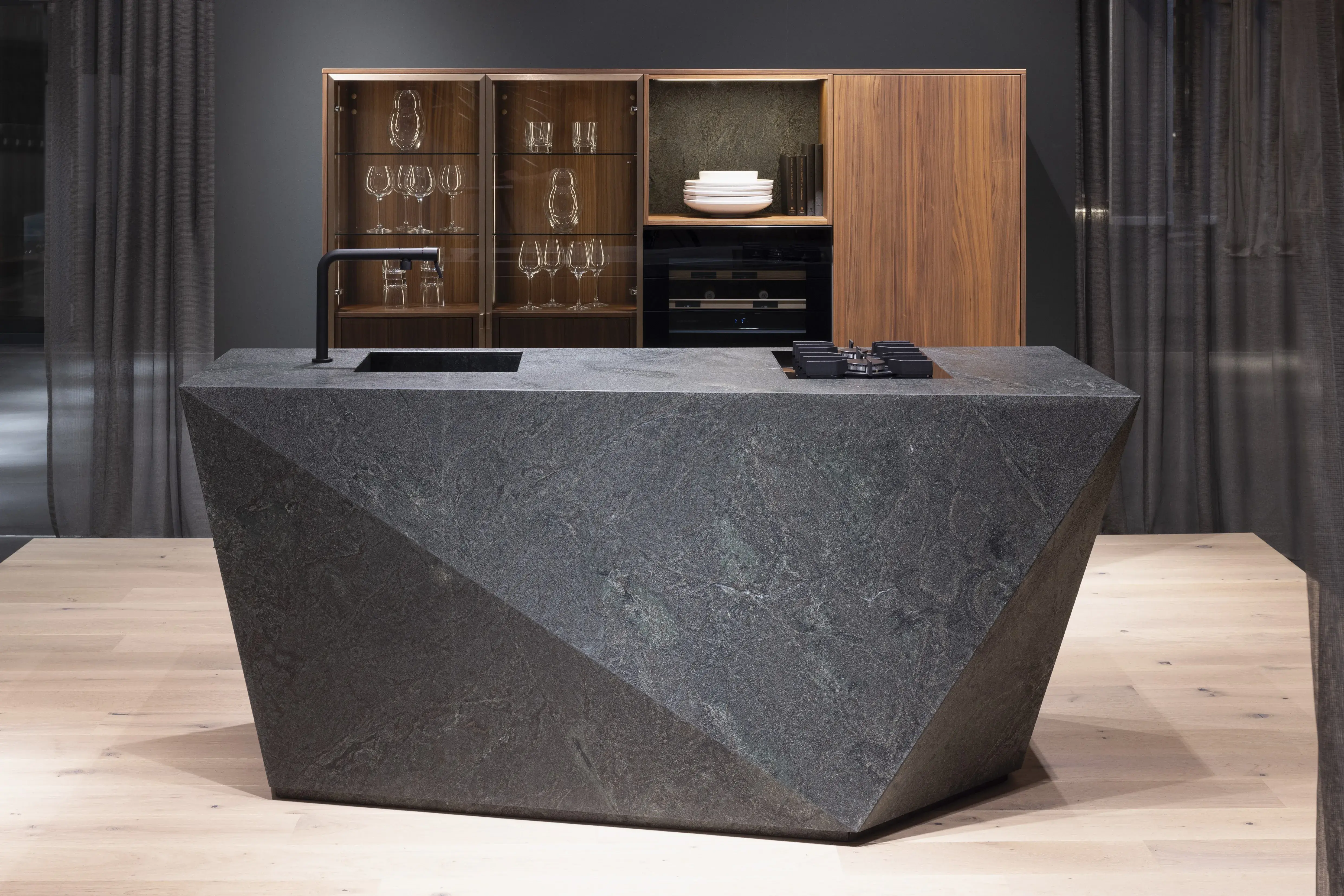 Block: Black Musk waterjet Front: AV 6085_GL Nussbaum-elegant Echtholzfurnier Korpus: 4077 Kristallweiß Griff: M745 Schwarz matt Geräte: Blaupunkt Armatur: Franke Spüle: Franke  Features: Polygon-Insellösung aus Naturstein, minimalistische Formgebung, beleuchtete Echtholzvitrinen, Einbau-Weinschrank